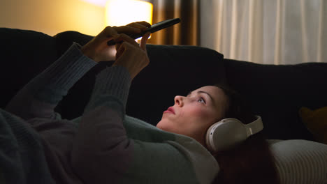 Mujer-Con-Auriculares-Inalámbricos-Tumbada-En-El-Sofá-De-Casa-Por-La-Noche-Transmitiendo-O-Mirando-Contenido-En-Línea-En-Un-Teléfono-Móvil